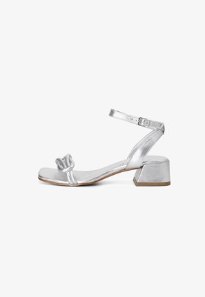 Sandal in metallo argentato con design anteriore annodato, cinturino alla caviglia regolabile e tacco quadrato, caratterizzato da una texture liscia e accenti minimi.