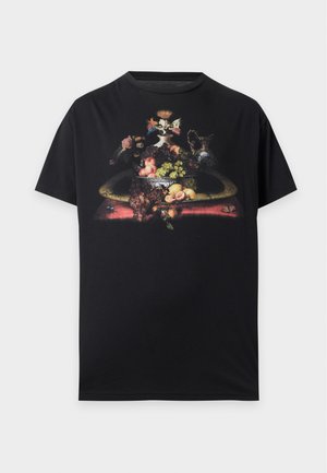Jeune homme debout, les mains dans les poches, portant un t-shirt noir à motif graphique, un pantalon cargo noir et des chaussures épaisses noires sur fond blanc.