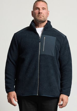 Homme portant une veste polaire bleu foncé avec un col montant et une poche poitrine zippée, debout devant un fond clair uni.