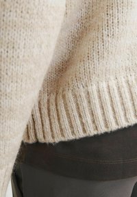 Beige knitted sweater with a ribbed hem, layered over a dark fabric. Soft texture, showcasing fine fibers and subtle variations in color.