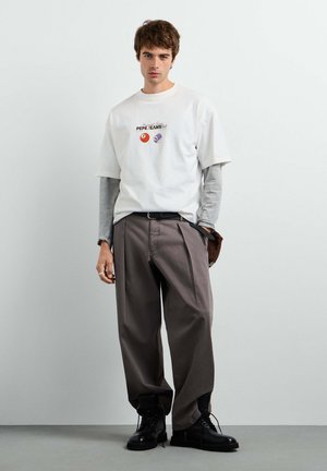 Giovane uomo che indossa una T-shirt grafica bianca su maniche lunghe grigie, pantaloni marroni larghi e stivali neri, in piedi contro una parete chiara e liscia.