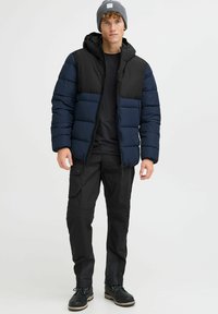 Polstret jakke i navy og sort, med hætte, lynlås lukning og quiltet design. Båret med sorte cargo bukser og sorte støvler.