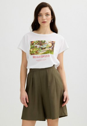 Joven mujer con cabello largo y castaño, que lleva una camiseta blanca con diseño gráfico y pantalones cortos plisados de talle alto en color verde oliva, posando frente a un fondo liso.