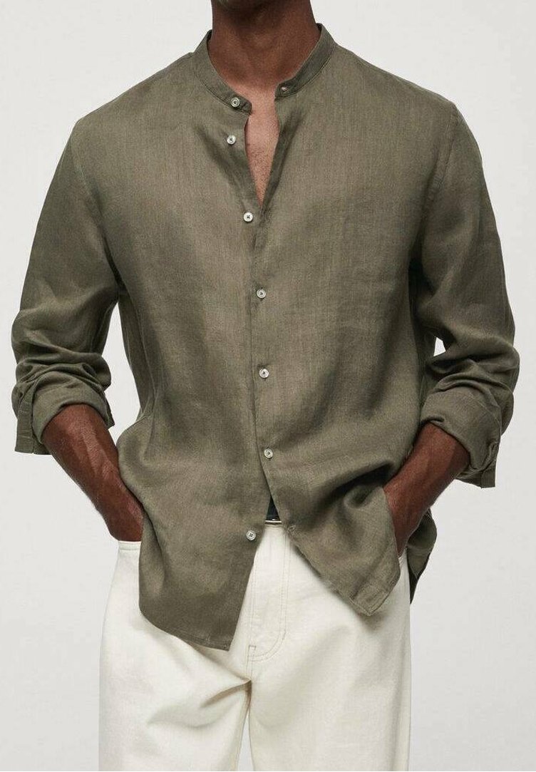 Homme portant une chemise boutonnée vert olive à manches longues avec col officier et manches retroussées, associée à un pantalon blanc. Mains dans les poches.