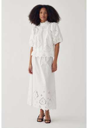Žena s kučeravými vlasmi v bielej blúzke s vyšívanými balónovými rukávmi a dlhou sukňou v rovnakej farbe, v kombinácii s čiernymi sandálmi s otvorenou špičkou, na jednofarebnom pozadí.
