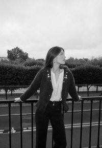 Grauer Cardigan mit floralem Stickmuster, weiße Bluse und dunkle Hose. Modell lehnt sich an ein Geländer mit grünem Laub im Hintergrund.