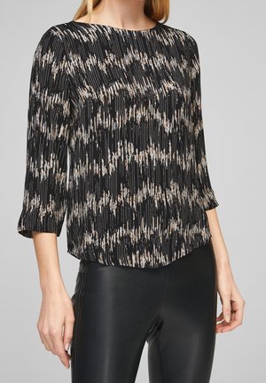 Femme portant un chemisier noir à manches longues avec des traits abstraits beige et blanc, associé à un pantalon noir aspect cuir.