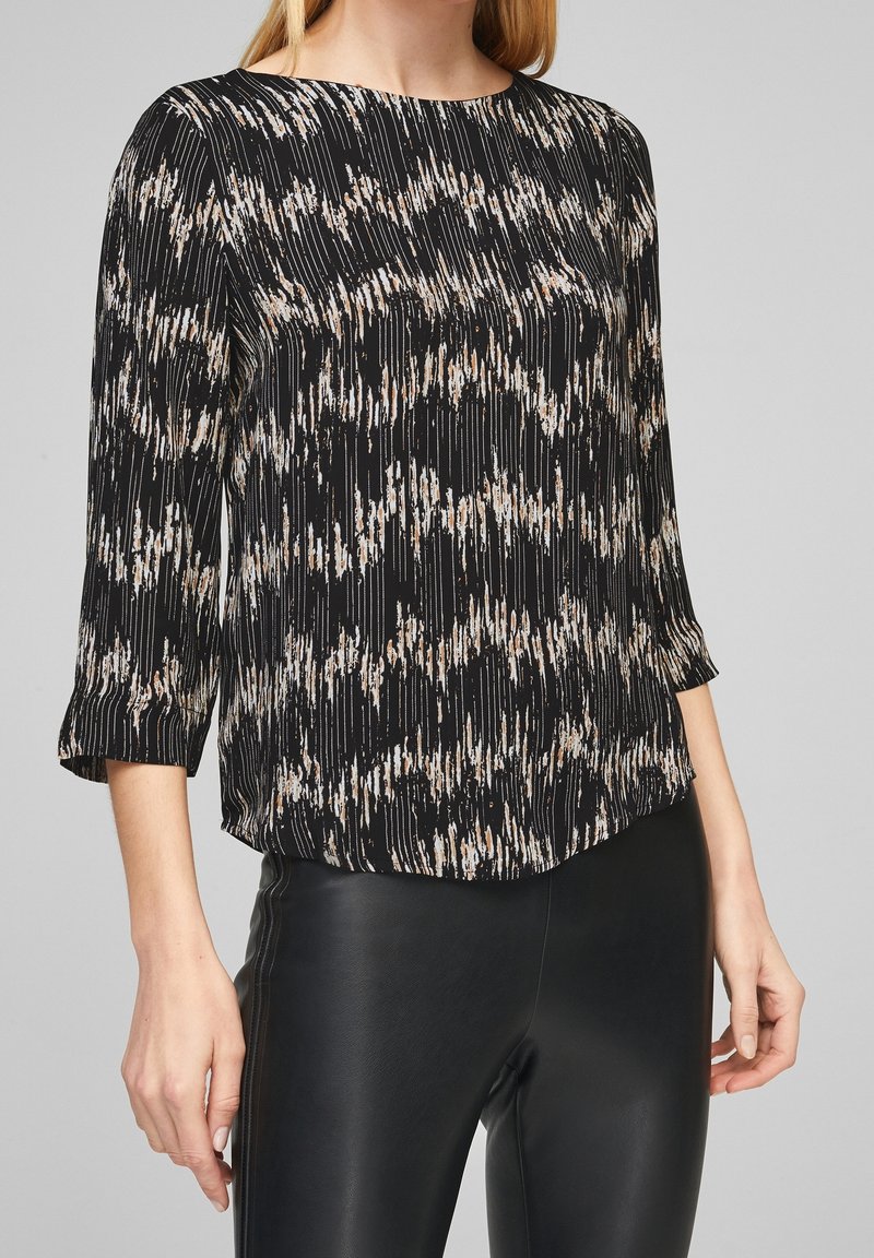 Femme portant un chemisier noir à manches longues avec des traits abstraits beige et blanc, associé à un pantalon noir aspect cuir.