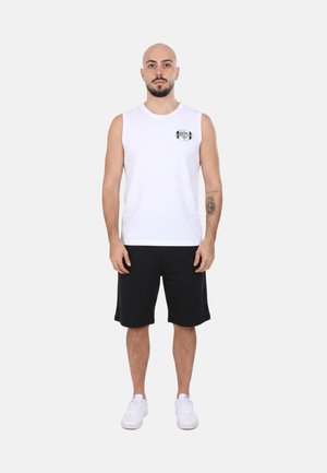 Uomo in piedi che indossa una canotta bianca con logo, pantaloncini neri, scarpe da ginnastica bianche e un tatuaggio sull'avambraccio sinistro, su sfondo neutro.