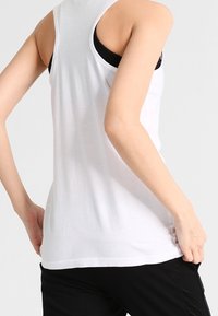 Biała bezrękawnikowa koszulka na ramiączkach wykonana z lekkiego materiału, z okrągłym dekoltem i luźnym krojem. Pod spodem widoczny czarny sportowy biustonosz.