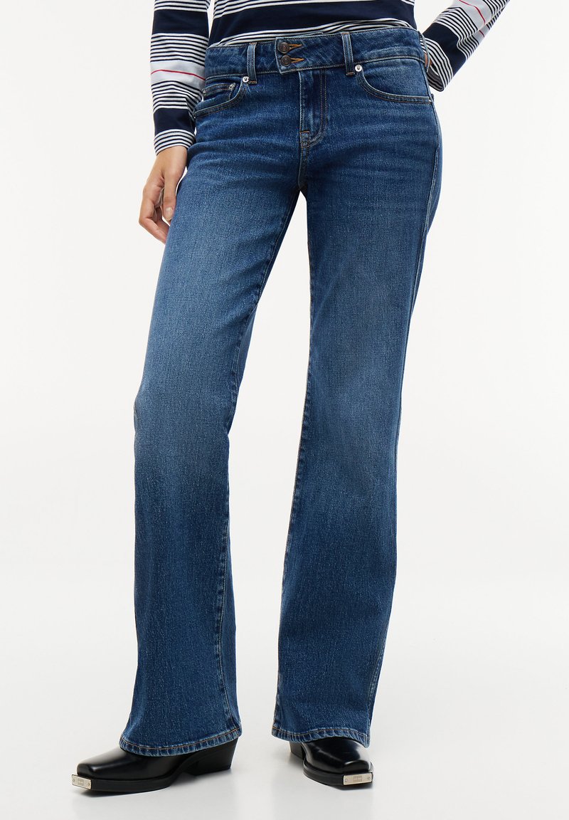 Γυναίκα με μπλε jeans bootcut, μαύρα παπούτσια με τακούνι και ριγέ μακρυμάνικη μπλούζα, όρθια με το ένα χέρι στην τσέπη.
