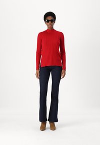 Rdeča rebrasta turtleneck pulover, dolge rokave, v kombinaciji s temnimi širokimi kavbojkami in rjavimi gležnjači. Dodatki vključujejo črne sončne očale.
