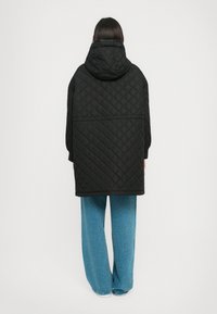 Persoon in een lange zwarte gewatteerde jas met capuchon en brede blauwe jeans, staande voor een simpele witte achtergrond.
