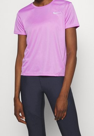 Persoon draagt een lichtpaarse korte mouw sportshirt met een wit Nike-logo en zwarte aansluitende sportleggings tegen een effen achtergrond.