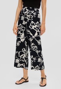 Femme portant un pantalon large et court noir avec un imprimé floral blanc et des sandales à talons noirs sur fond gris uni.