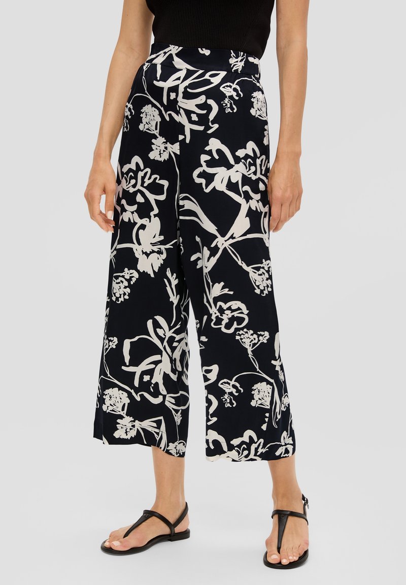 Femme portant un pantalon large et court noir avec un imprimé floral blanc et des sandales à talons noirs sur fond gris uni.