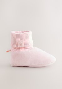 Chaussons en peluche roses avec un bord côtelé et une étiquette portant un logo fleur, bout arrondi, et texture douce pour le confort.
