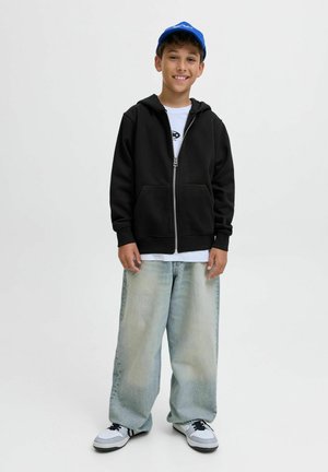 Haut à capuche noir zippé avec poches avant, t-shirt graphique blanc, jeans larges bleu clair, et casquette bleue, associés à des baskets noires et blanches.