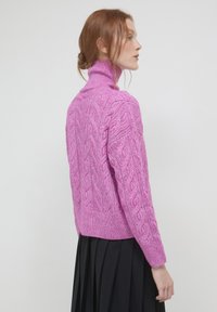 Frau mit roten Haaren im Dutt, die einen rosa Rollkragenpullover aus Kabelstrick und einen schwarzen Plisseerock trägt, seitlich vor einem einfarbigen Hintergrund stehend.