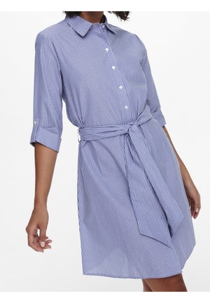 Femme portant une robe chemise à rayures verticales bleues et blanches avec des manches retroussées et une ceinture nouée à la taille, debout devant un fond blanc.