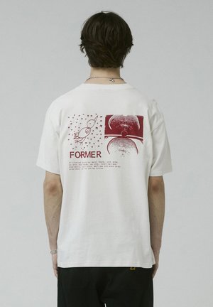Persoon die een wit T-shirt draagt met een rode vogel, abstracte cirkelvormige ontwerpen en het woord "FORMER" op de achterkant gedrukt.