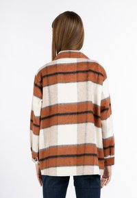 Flanellen shirt in oranje, crème en zwarte geruite patroon. Heeft een kraag, lange mouwen en knopenmanchetten. Zachte textuur.