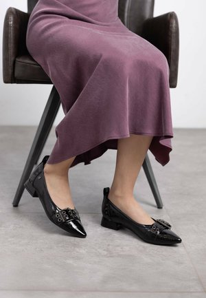 Bailarinas de charol negro con punta afilada, que cuentan con una hebilla decorativa y acentos de tachuelas, sobre una persona sentada en una silla.