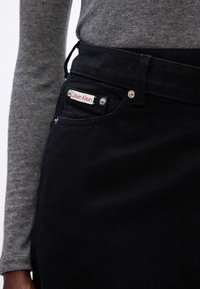 Jeans in denim nero con cuciture visibili, un bottone argentato e un'etichetta "Calvin Klein" rossa e bianca sulla tasca anteriore. Manica di t-shirt grigia.