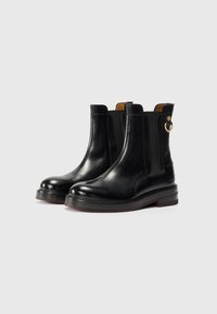 Botas de couro pretas até ao tornozelo com painéis laterais elásticos, ponta arredondada e sola de 3 cm de espessura. Apresenta um detalhe em metal dourado na parte de trás.