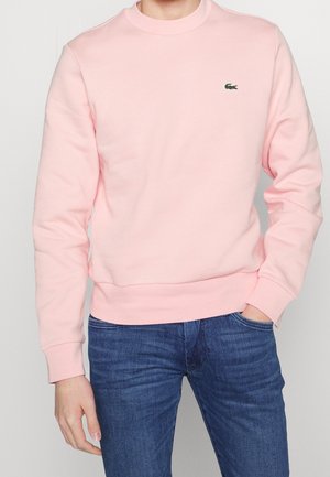 Mies, joka pukeutunut vaaleanpunaiseen pyöreäkaulaiseen collegepaitaan, jossa pieni vihreä krokotiililogo rinnassa, ja sinisiin farkkuihin, käsi taskussa.