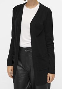 Cardigan en maille noire avec ouverture devant, manches longues et deux poches avant. Porté sur un haut blanc, associé à un pantalon noir. Texture lisse.