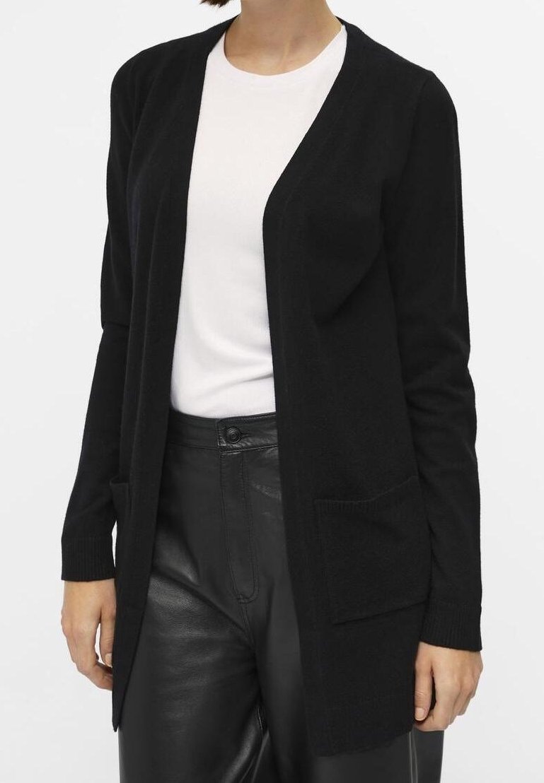 Cardigan en maille noire avec ouverture devant, manches longues et deux poches avant. Porté sur un haut blanc, associé à un pantalon noir. Texture lisse.