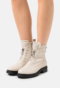 Beige ankle boots made of leather, featuring prominent studded accents, dual side zippers, and a black textured rubber sole.