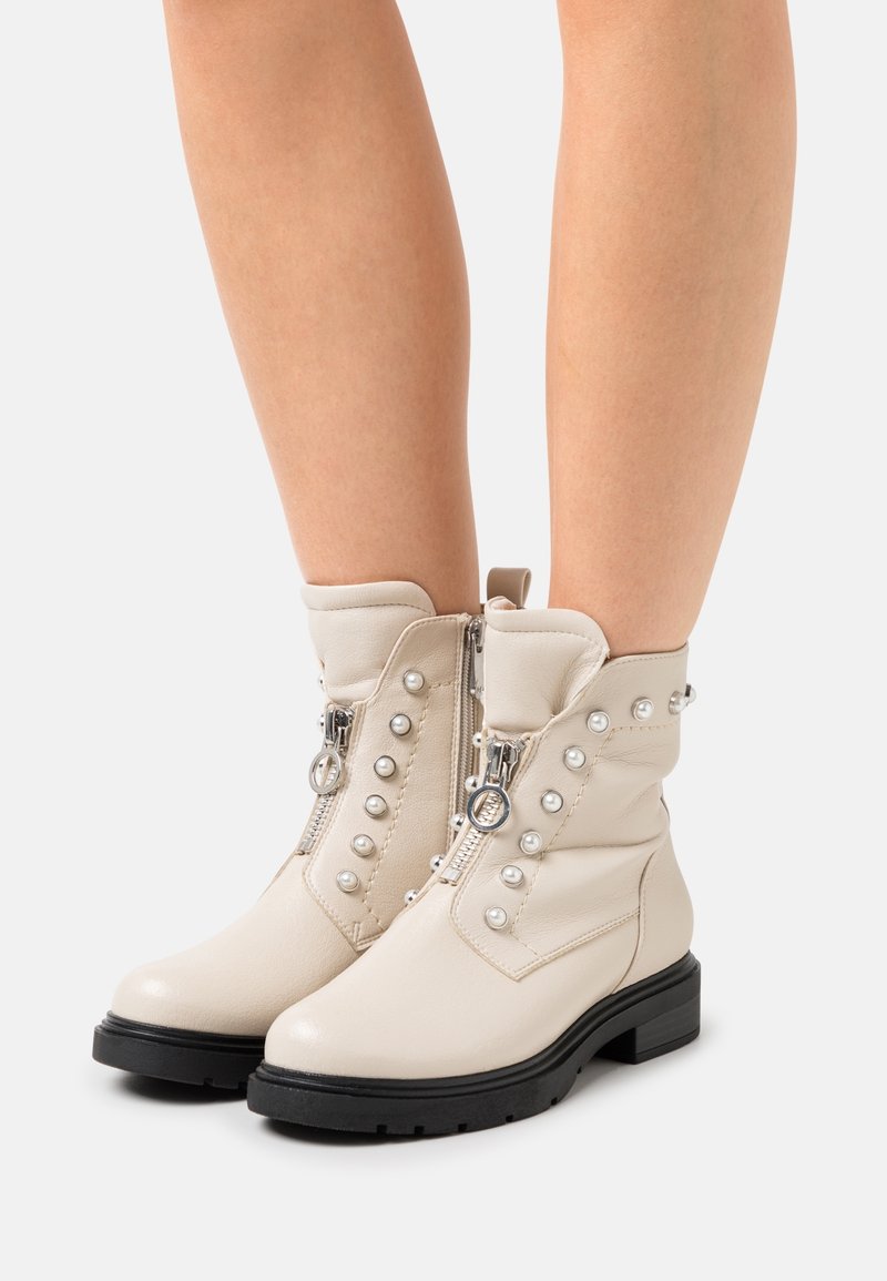 Beige ankle boots made of leather, featuring prominent studded accents, dual side zippers, and a black textured rubber sole.