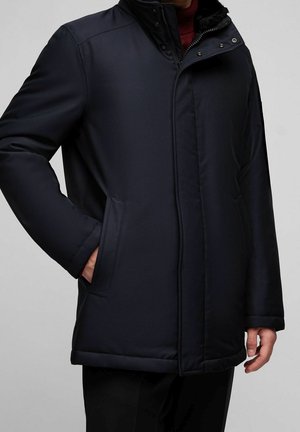 Mann trägt einen dunkelblauen, gesteppter Wintermantel mit Seitentaschen, Reißverschluss und Knopfverschluss und hohem Kragen über einem roten Pullover.