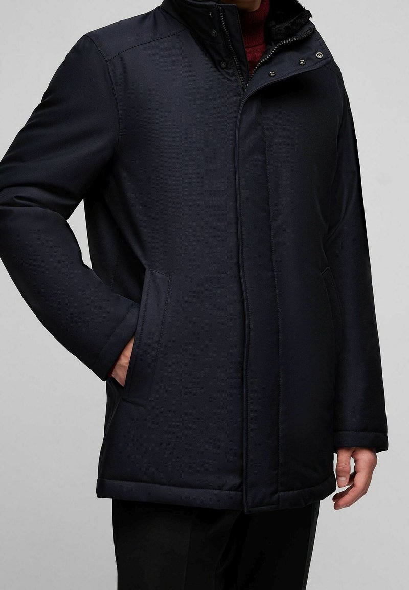 Homme portant un manteau d'hiver matelassé bleu marine foncé avec poches latérales, fermeture éclair et boutons, et un col montant par-dessus un pull rouge.