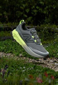 Γκρι συνθετικό αθλητικό παπούτσι με υφή στην επάνω επιφάνεια, αντανακλαστικές λεπτομέρειες και φωτεινή κίτρινη σόλα, εμφανιζόμενο μπροστά σε φόντο με γρασίδι.