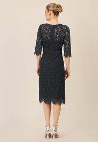 Vestido de encaje con mangas tres cuartos, color verde azulado oscuro, corsé ajustado y dobladillo en ondas. Zapatos plateados completan el look.