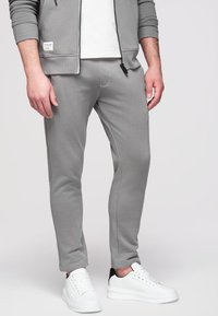 Homme portant un pantalon de jogging gris, des baskets blanches avec un détail noir au talon et une veste zippée grise par-dessus une chemise blanche, debout sur un fond blanc.