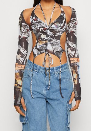 Primo piano di una persona che indossa un top a collo alto con stampa astratta e ritagli, maniche lunghe staccate e pantaloni cargo larghi in denim blu.