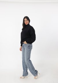 Zwarte hoge hals trui met ribbels aan de mouwen, gecombineerd met lichtblauwe high-waisted jeans en witte hakken met een diamanten patroon.