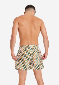 Costumi da bagno bianchi con un motivo ondulato arancione e verde, vita elasticizzata e tasche laterali. Il tessuto sembra leggero e liscio.