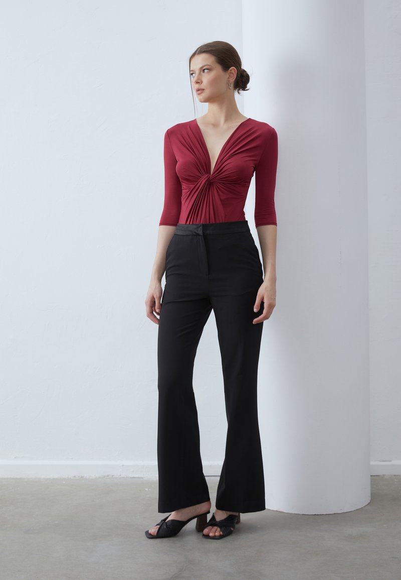 Top aderente bordeaux con maniche a tre quarti, caratterizzato da un nodo centrale. Abbinato a pantaloni neri a vita alta e sandali neri con tacchi.