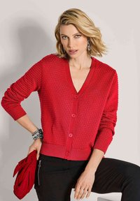 Frau mit blonden Haaren, die einen strukturierten roten Knopfcardigan, schwarze Hosen, silberne Ohrringe und Armbänder trägt und eine rote Clutch-Tasche hält.