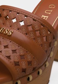 Καφέ δερμάτινο πλατφόρμα σανδάλι με ανοίγματα, χρυσά καρφιά κατά μήκος της άκρης της σόλας και χρυσό λογότυπο στο λουράκι.