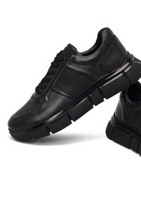 Des baskets en cuir noir présentent une texture lisse, un bout rond, des lacets plats et une semelle en caoutchouc épaisse avec des motifs proéminents et des panneaux d'accent.