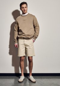 Sudadera beige con cuello redondo y un logo sutil, combinada con pantalones cortos beige claros. El calzado presenta tonos blancos con acentos marrón claro. Tejido texturizado.