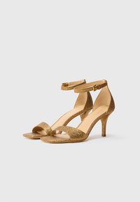 Tacones abiertos de oro con purpurina, tiras en el tobillo y tacones delgados de altura media, exhibidos sobre un fondo blanco.