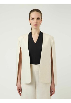 Kvinne med tilbakestrøket hår har på seg kremfarget kappeblazer over svart V-hals-topp og matchende kremfargede bukser mot ensfarget bakgrunn.