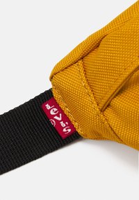 Angolo di una borsa di tessuto gialla con cuciture visibili, attaccata a una tracolla nera, che presenta un piccolo tag rosso con testo bianco su sfondo bianco.
